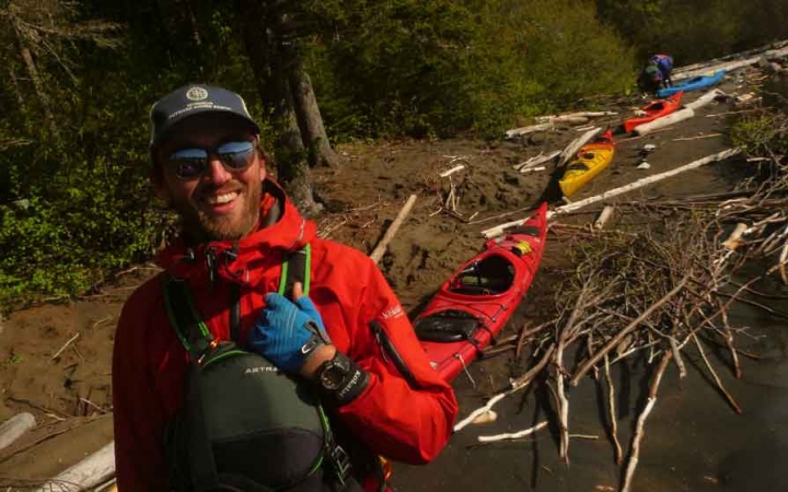 a student smiles at the camera with kayaks resting on the shore in the background